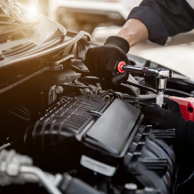 A mechanic wearing black gloves uses a ratchet wrench on an engine under a car's open hood.