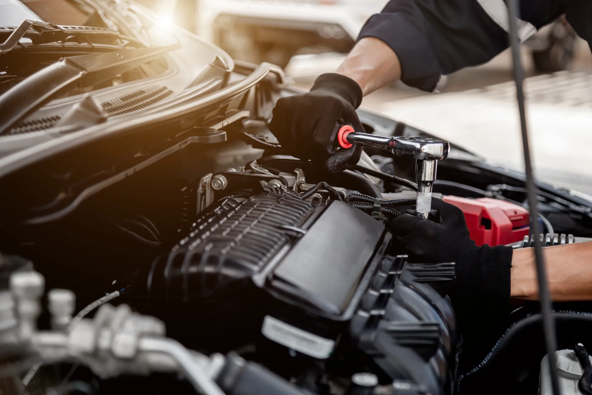 A mechanic wearing black gloves uses a ratchet wrench on an engine under a car's open hood.