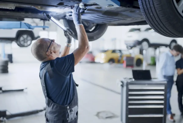 A mechanic in overalls and safety glasses works underneath a car raised on a lift in a busy auto repair shop.