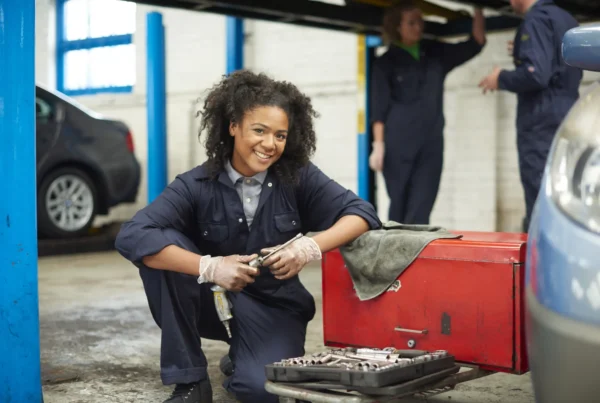 A smiling female mechanic in navy coveralls kneels with an air ratchet next to a red tool chest inside an auto repair garage while two technicians work under a lifted car behind her.