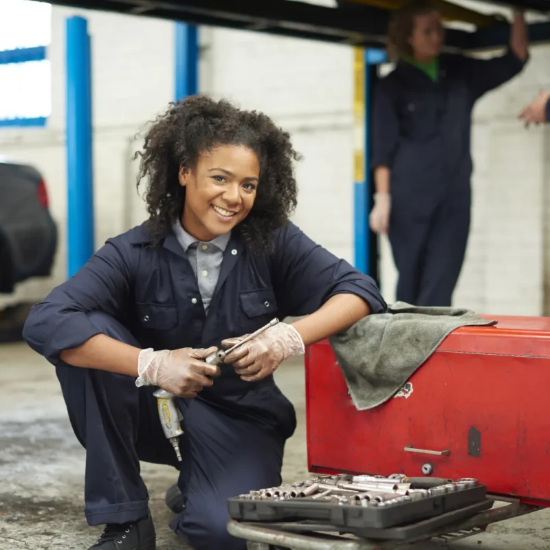A smiling female mechanic in navy coveralls kneels with an air ratchet next to a red tool chest inside an auto repair garage while two technicians work under a lifted car behind her.